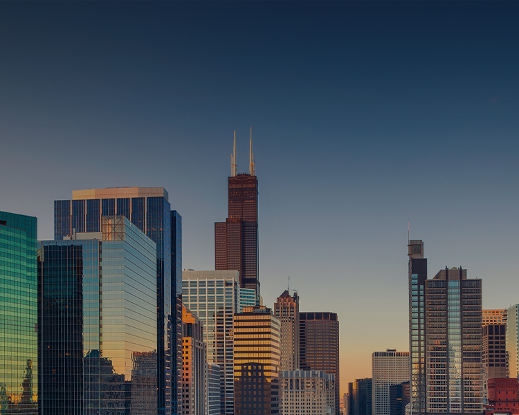 A panoramic view of a city skyline during sunset, showcasing modern skyscrapers with the prominent tower having a reddish-brown hue. The buildings reflect the golden light of the setting sun against a clear blue sky.
