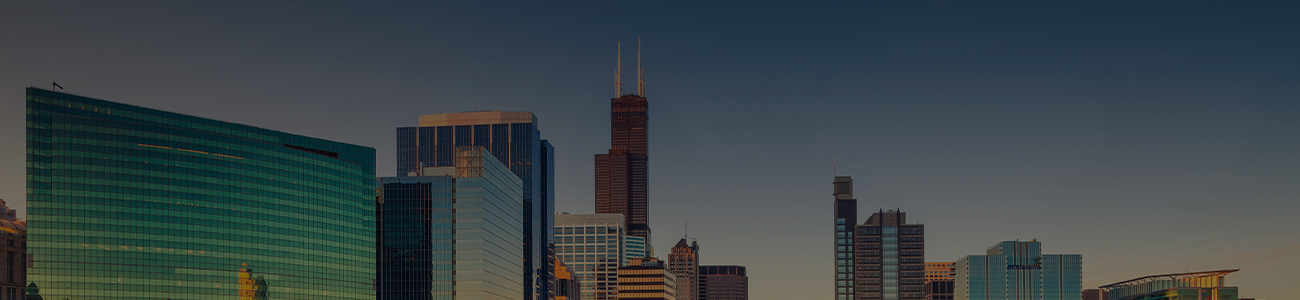 A panoramic view of a city skyline during sunset, showcasing modern skyscrapers with the prominent tower having a reddish-brown hue. The buildings reflect the golden light of the setting sun against a clear blue sky.